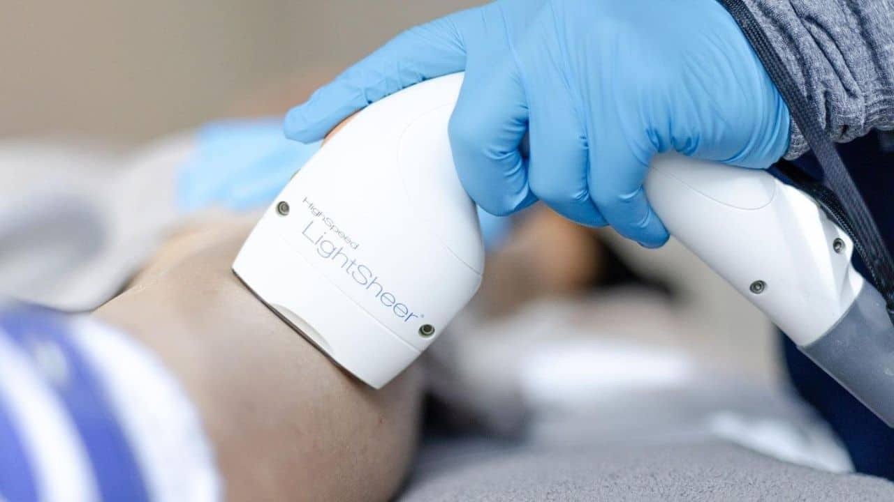 Laser hair removal device applied to a person’s leg by a technician wearing blue gloves in a clinical setting