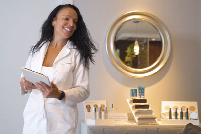 Smiling skincare professional in a white coat holding a tablet, standing beside a display of skincare products in a clinic setting.